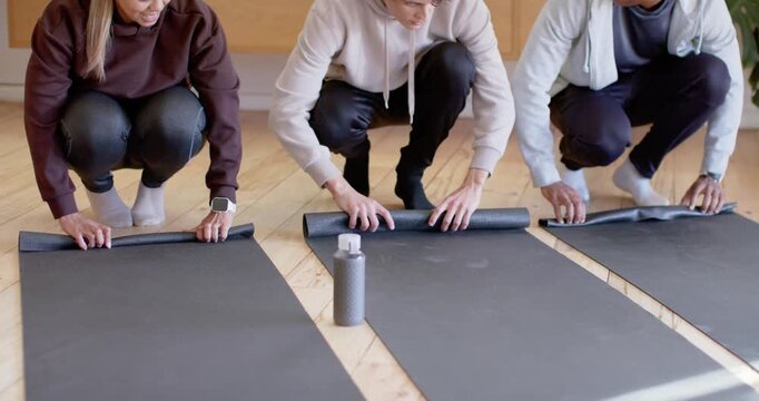 Diverse trio squatting center leading rolling black mats while prepping workout near gray bottle