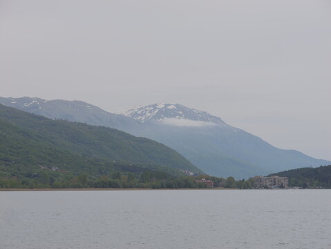 View of the calm waters of Lake Ohrid with snow-capped mountains rising in the background under an overcast sky in Ohrid, Municipality of Ohrid, North Macedonia.