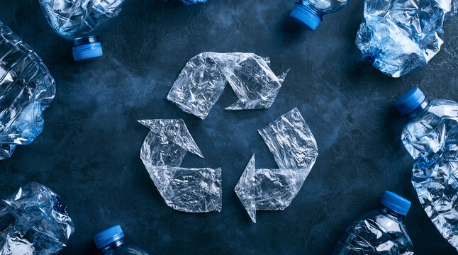 A recycle symbol made from crumpled clear plastic is surrounded by blue-capped bottles on a dark background. It illustrates environmental conservation and plastic recycling.