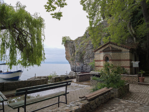 View of the small stone chapel near rocky cliffs and Lake Ohrid, with a wooden boat moored at the shore and a park bench in the foreground, Ohrid, Municipality of Ohrid, North Macedonia.