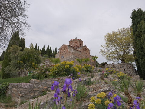 View of the historic Church of St. John at Kaneo surrounded by stone walls, blooming purple irises, and cypress trees in Ohrid, Municipality of Ohrid, North Macedonia.