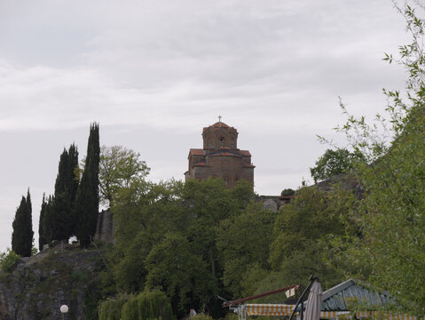 View of the Church of St. John at Kaneo perched on a cliff overlooking the lake, surrounded by lush trees and tall cypress trees under a cloudy sky in Ohrid, Municipality of Ohrid, North Macedonia.