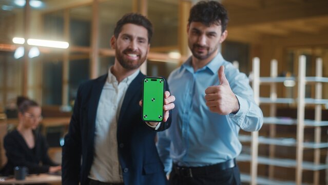 Two Caucasian business men smiling happy showing mobile phone with green screen demonstrate thumb up like gesture cheerful positive businessmen male partners recommending gadget online app in office