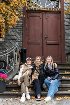 Three smiling women sit on stone steps before historic doorway during autumn trip in Harz, Germany. Candid travel portrait shows friendship, tourism, leisure and warm outdoor seasonal atmosphere