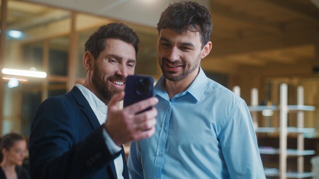Two Caucasian business men looking in mobile phone screen watching online content smiling laughing in office. Cheerful businessmen male colleagues with device watch social media video smile together