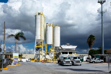 Fototapeta premium Industrial concrete batching plant factory with tall white storage silos and cement mixer delivery trucks