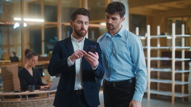 Two concentrated Caucasian business men serious looking in mobile phone scrolling social media online internet service businessmen male partners workers look in gadget device app teamwork in office