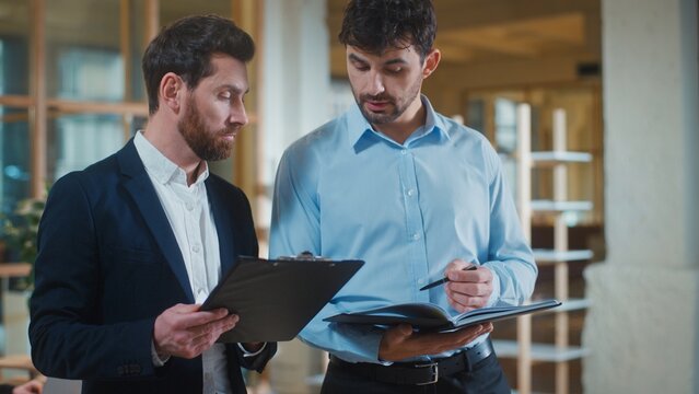 Two Caucasian business men comparing data with documents talking discussing stats in office. Focused businessmen executives colleagues talk discussing work results with papers male partners teamwork