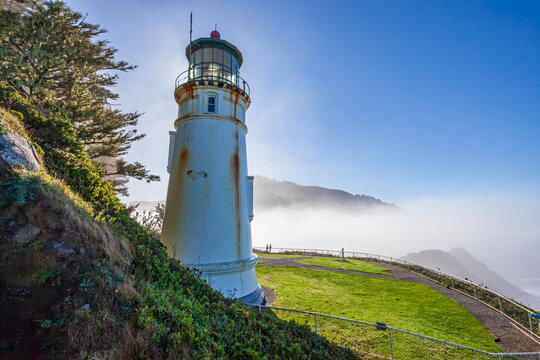 Beautiful scenic view of a historic white lighthouse on a green cliff next to a misty ocean at sunrise