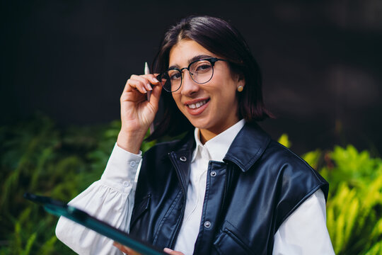Hispanic woman copywriter with electronic stylus tablet posing looking at camera with positive attitude. Spanish female content creator managing freelance social media network update in park.