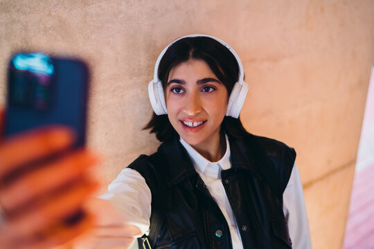 Spanish female selfie look directly at camera smartphone screen text chatting. Happy hispanic woman student video call application communication posing nearby copy space texturized wall layout.