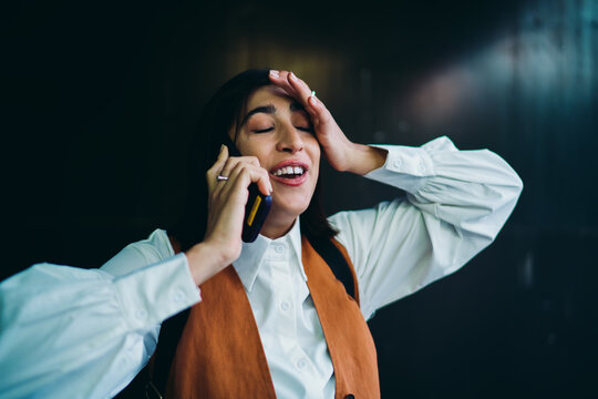 Disappointed latin female student gesturing during cellular conversation network problem nearby dark wall background. Upset hispanic woman holding smartphone touching head with closed eyes.