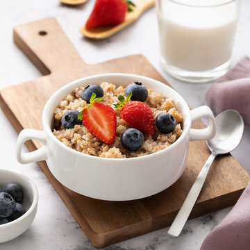 Buckwheat flakes with strawberry and blueberries in white bowl on wooden tray, glass of milk on table, healthy breakfast