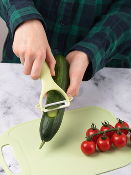 Teenager peeling cucumber with vegetable peeler in kitchen, close-up of hands, cherry tomatoes on cutting board nearby