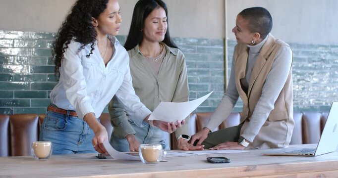 Diverse female team receiving printouts leaning over table at cafe planning project with tablet
