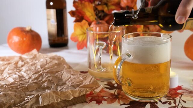 Person pouring foamy beer into a glass mug on a table with an autumn tablecloth, illustrating a cozy home Oktoberfest atmosphere.