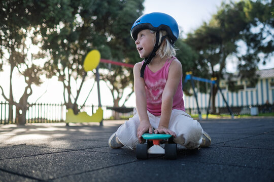 Cute Little Girl in Blue Helmet Sitting on Penny Board at Playground Outdoors