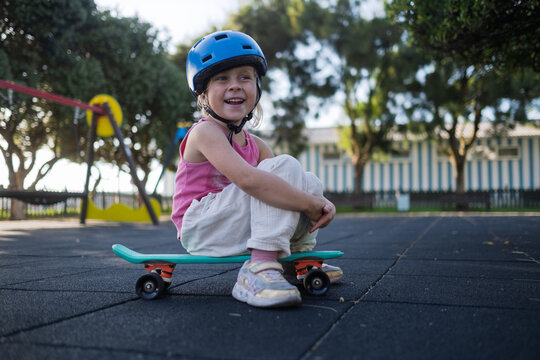 Cute Little Girl in Blue Helmet Sitting on Penny Board at Playground Outdoors