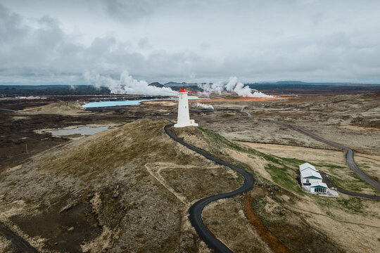Aerial view of the Reykjanesviti lighthouse standing on a hill with geothermal steam rising in the background and a winding road leading to the structure in Reykjavik, Reykjavikurborg, Iceland.