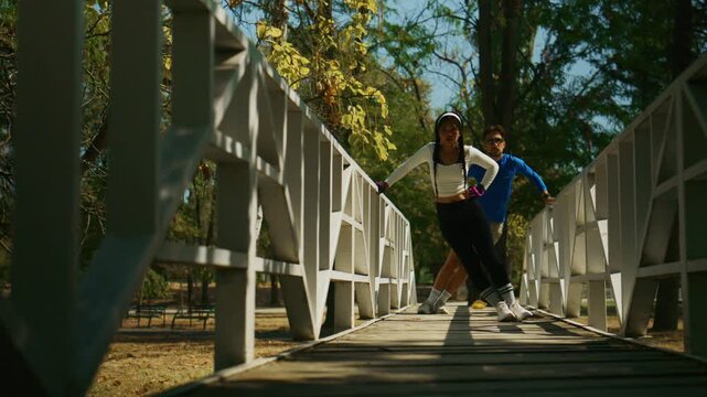 Two young people lean playfully on a white wooden bridge in a sunny park. They enjoy a relaxed and active moment outdoors among trees and fallen leaves.