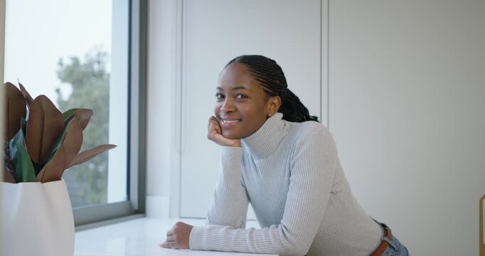 African woman leaning on window ledge with planter, noticing camera and laughing in gray sweater