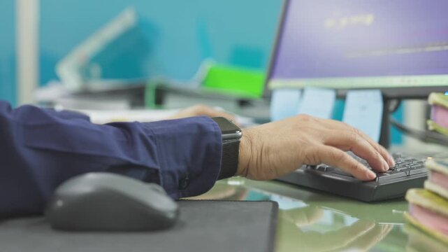 Focused male programmer types data, operating keyboard beside computer mouse, reviewing desktop monitor screen, managing daily digital workflow inside corporate tech office workplace setting.