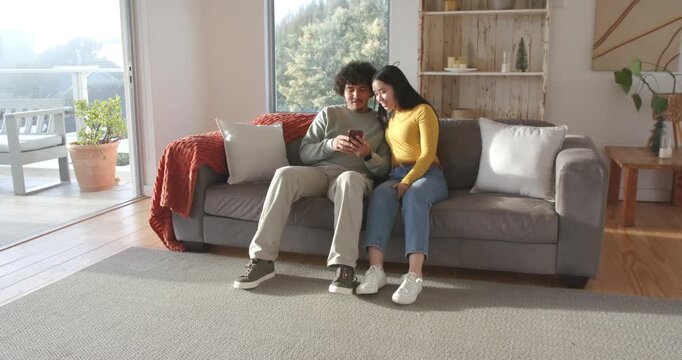 African American man, Asian woman on sofa viewing smartphone, reacting, standing, dancing at home