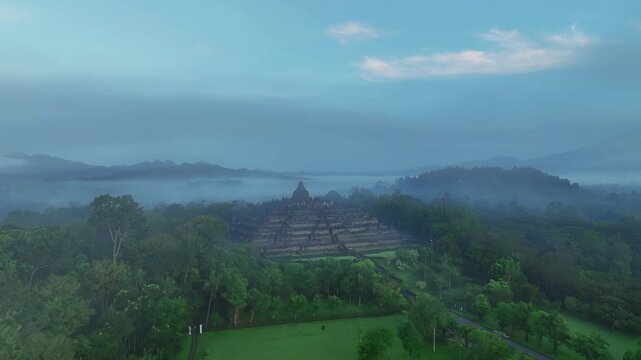 Aerial view of the ancient Borobudur temple surrounded by lush tropical trees and misty mountains in Borobudur, Central Java, Indonesia.