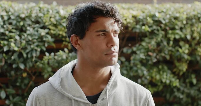 African American man standing by leafy hedge wearing gray hoodie, turning to sunlight and smiling