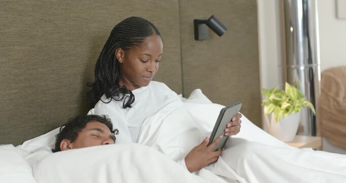 African couple lying in bed while man sleeping and woman holding tablet, reading to unwind