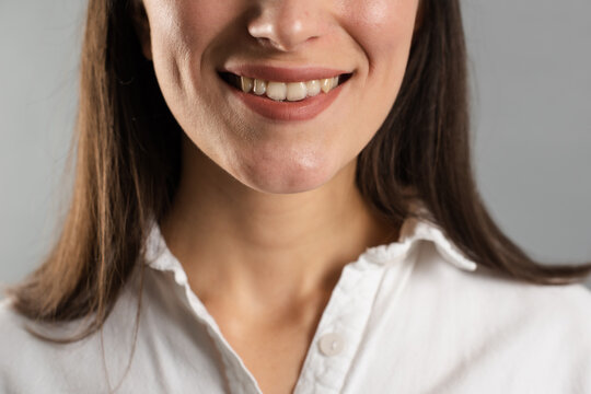 Crop view of smiling young woman face with healthy teeth and dimples wearing white shirt on gray background