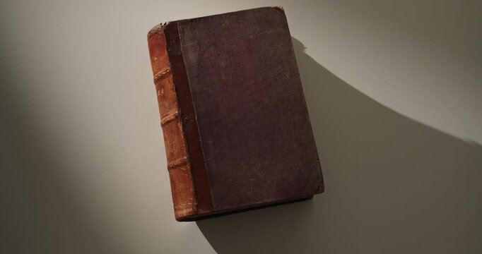 Cinematic orbit around an old vintage book with a worn leather spine resting on a white surface, illuminated by atmospheric local light and casting long dramatic shadows. Education, history, knowledge