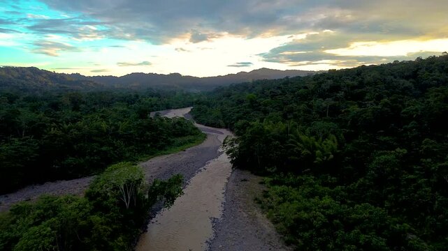 Aerial view following a winding muddy river between dense tropical rainforest and forested hills beneath a glowing evening sky. The tranquil wilderness represents freshwater habitat, biodiversity and 