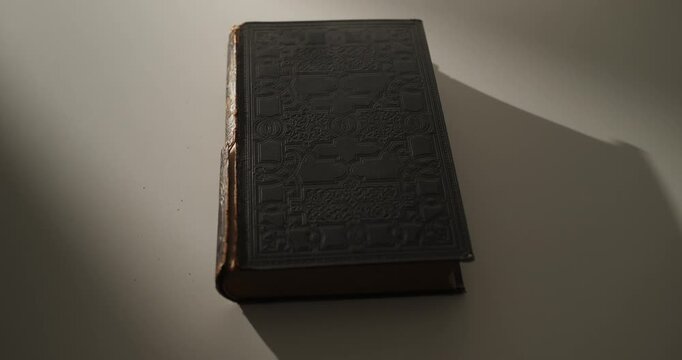 Cinematic orbit around an old vintage book with a worn leather spine resting on a white surface, illuminated by atmospheric local light and casting long dramatic shadows. Education, history, knowledge