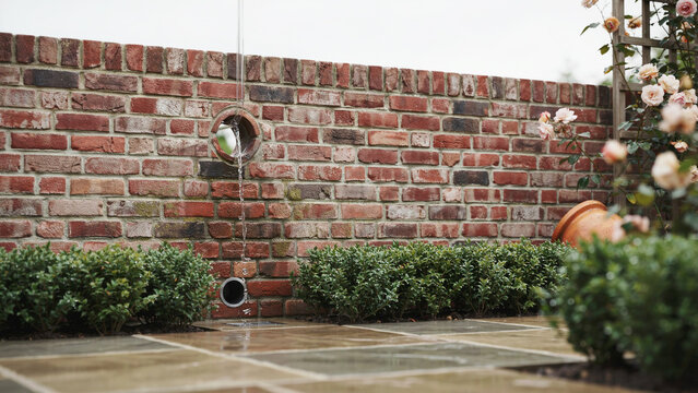 Water fountain feature embedded in rustic brick wall surrounded by lush garden plants and flowers