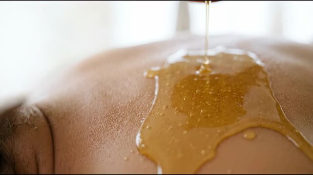Close-up of golden honey pouring onto a bare back from a terracotta pot. Natural skincare therapy and spa treatment concept. Macro detail of sweet liquid flowing