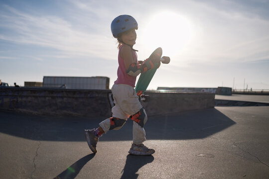 Happy Little Girl in Blue Helmet Carrying Penny Board at Skatepark in Sunlight