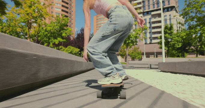 Rear tracking shot follows young woman crouching on longboard through modern city park with dynamic confidence. Skate culture, urban movement and youth freedom concept.