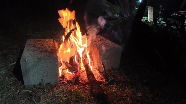 Static view of bright fire flames and wood logs burning in an improvised cinder block fire pit outdoors at night.