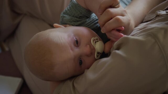 Caucasian baby sucking pacifier and holding mother fingers while lying down. Clip suitable for parenting campaigns, pediatric healthcare, and infant product advertising