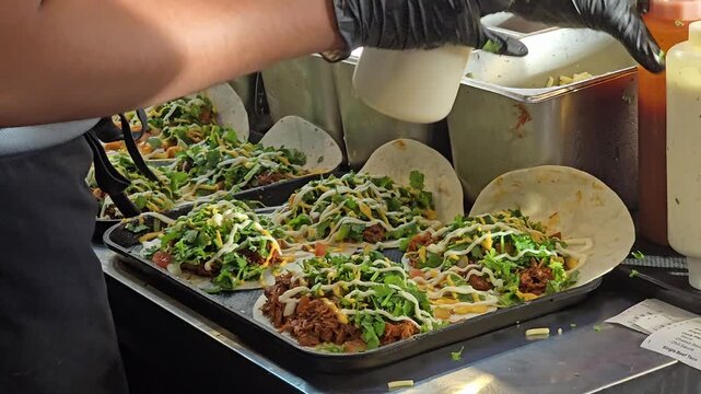 Close-up of a chef wearing black gloves drizzling sauce over fresh Mexican tacos filled with seasoned meat, cheese, and shredded lettuce.