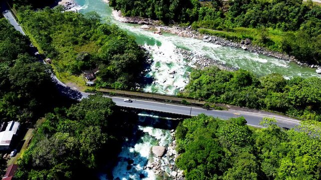 Top-down aerial view of cars travelling across a road bridge above a rocky whitewater river surrounded by lush vegetation. The scene combines transport infrastructure, mobility and natural landscape.