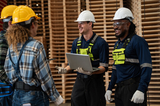 A diverse team of workers gathers inside a large warehouse stocked with wooden pallets, sharing a moment of strong teamwork and collaboration while reviewing project details on a laptop.