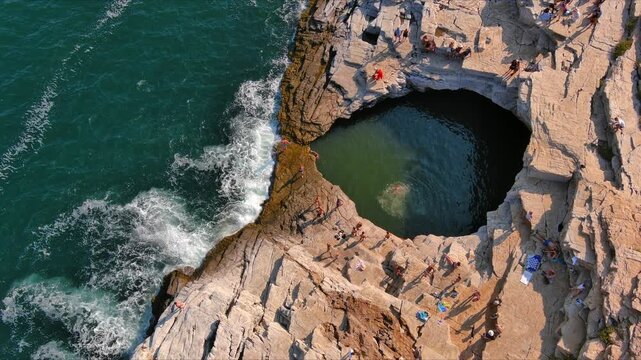 Aerial view of the natural rock pool Giola with people swimming in the turquoise water surrounded by rugged cliffs along the coastline of Thasos, Greece.