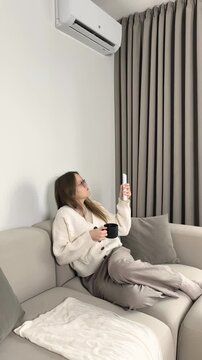 Air conditioner remote control used by woman sitting on sofa with hot tea cup to heat room. Female person holding warm mug and adjusting domestic temperature during cold winter day. Chilly