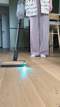 Cordless vacuum cleaner brush nozzle clearing dust from engineered wood floor surface at home. Modern household appliance with blue light illumination used for domestic floor care routine.