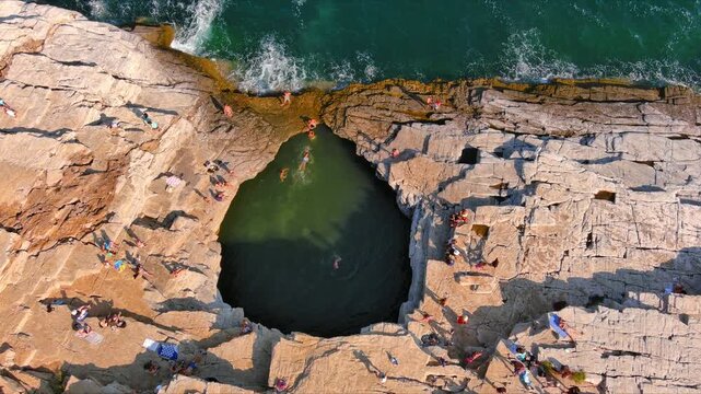 Aerial view of people swimming in the Giola natural rock pool surrounded by rugged cliffs and turquoise sea in Thasos, Greece.
