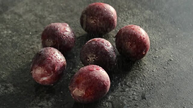 Fresh ripe plums are glistening with water droplets and arranged in a circular pattern on a dark textured surface.  water droplets fallen on plums