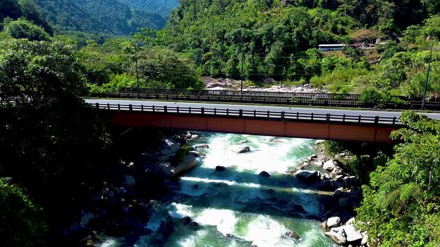 Cargo truck travels across a road bridge above a fast-flowing turquoise river surrounded by lush tropical mountains. The scene highlights logistics, transport infrastructure, regional connectivity and