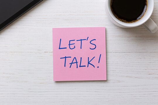 Note with handwritten lets talk message on a pink sticky note at a white wooden desk with coffee